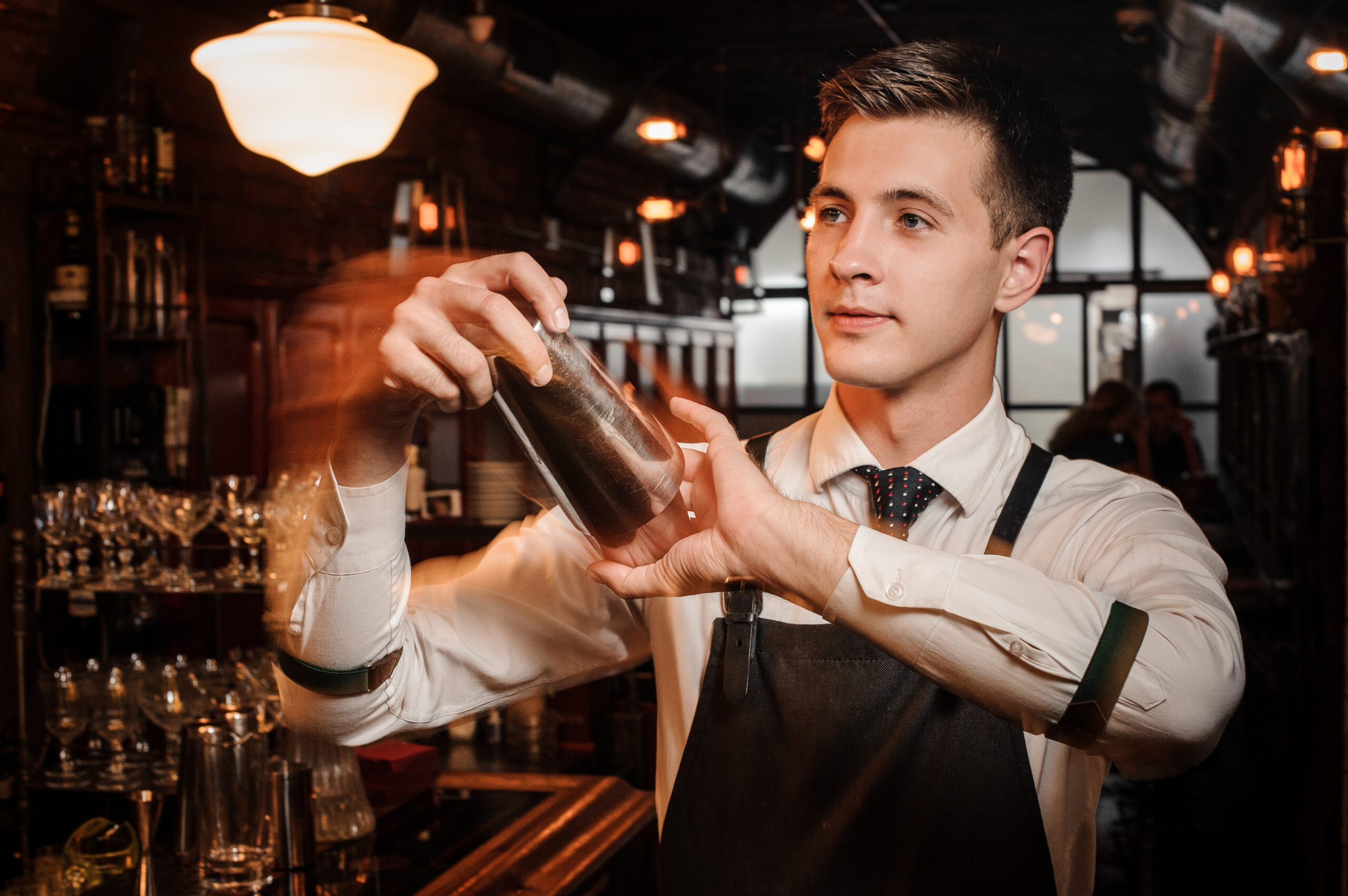 A bartender mid-shake with a metal cocktail shaker behind a busy bar, motion blur in the hands and warm ambient lighting