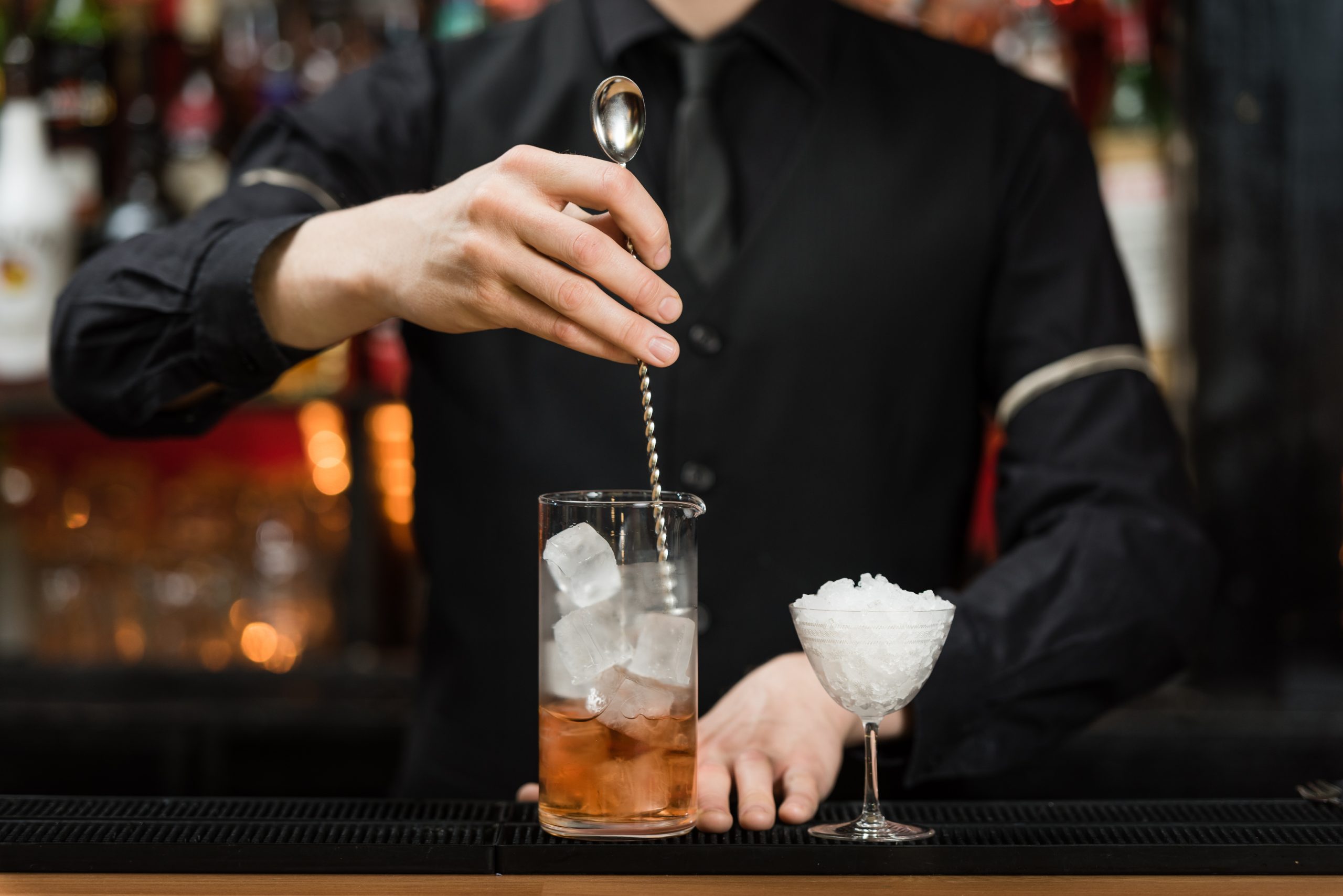 A bartender stirring a drink in a mixing glass behind a bar, with an empty coupe glass on the counter