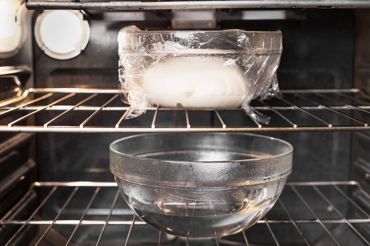 A covered bowl of bread dough resting on an oven rack with the oven light on, creating a warm proofing environment