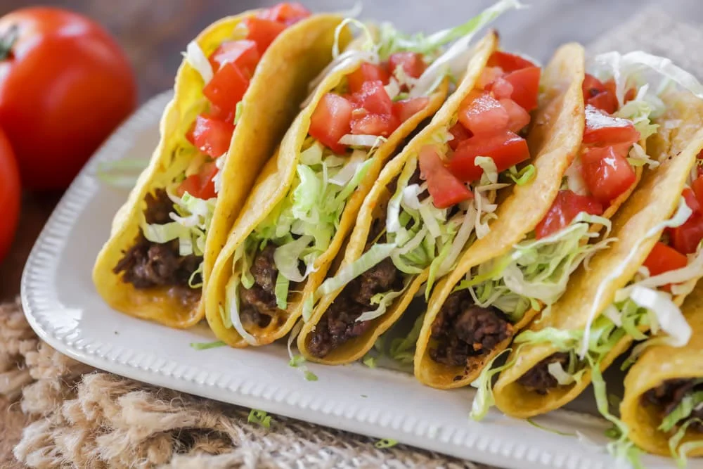 A plate of corn tortilla tacos with grilled meat, onions, cilantro, and lime wedges on a wooden table at a casual restaurant