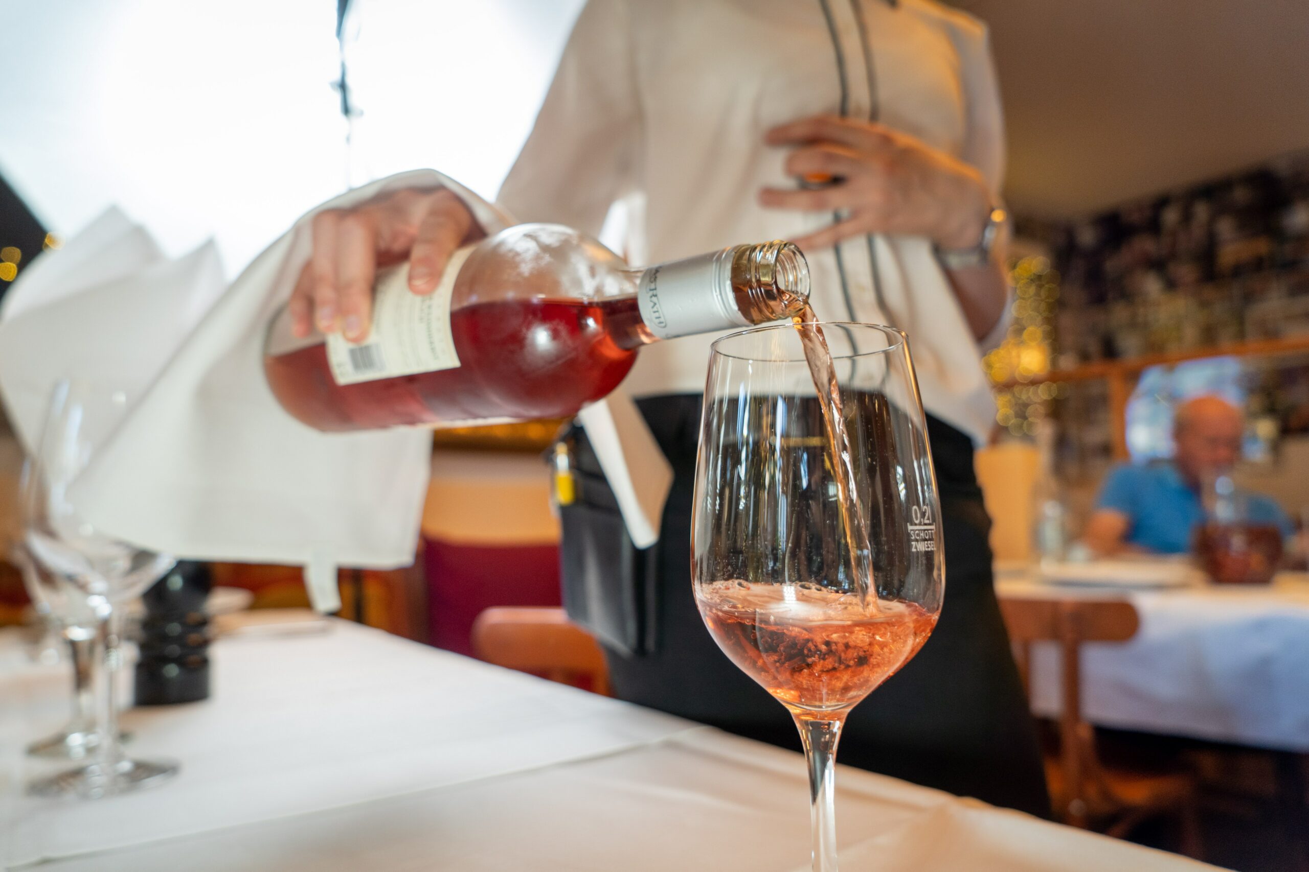 A sommelier presenting a wine bottle tableside in a softly lit dining room with guests seated in the background