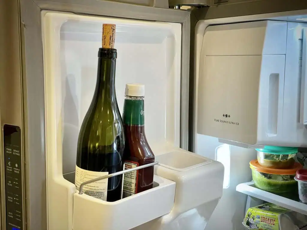 An open bottle of red wine with the cork reinserted, standing upright on a refrigerator shelf next to everyday groceries